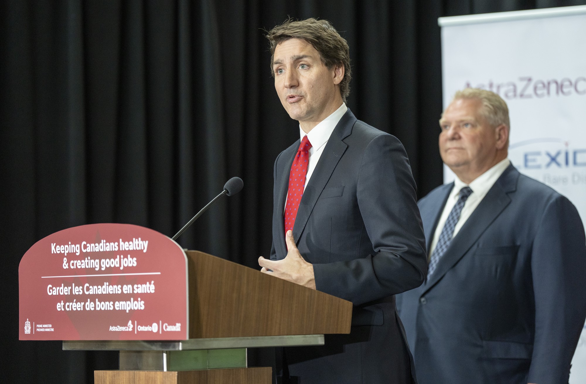 Justin Trudeau stands at a podium speaking as a person in a suit stands behind in the background.