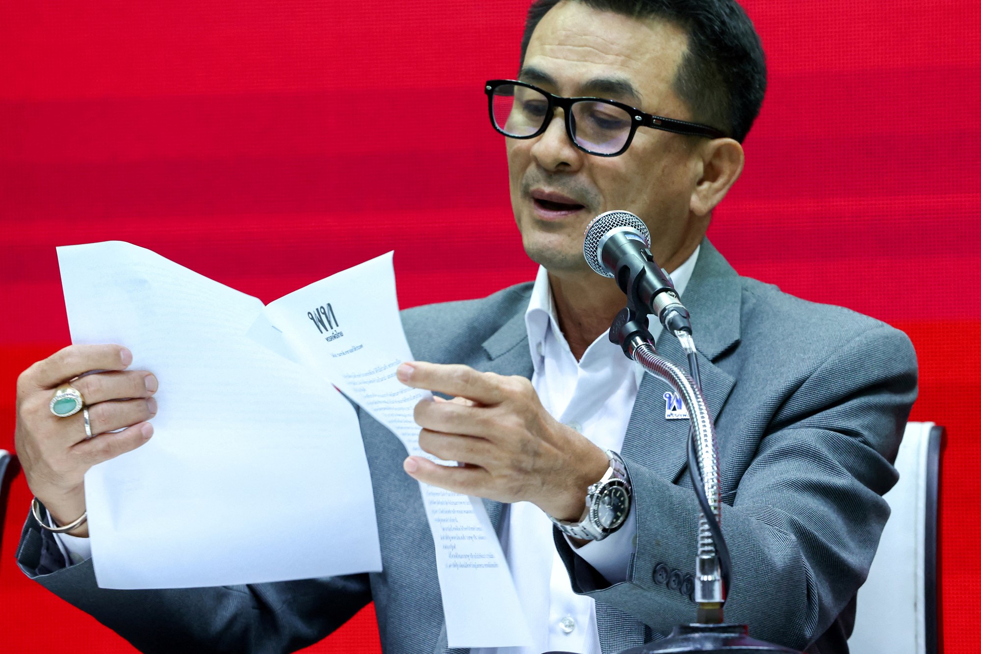 A man in a grey suit, glasses holds a piece of paper and speaks into a mic