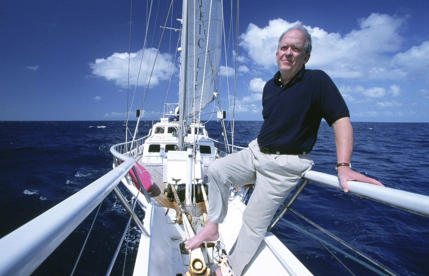 Roger Payne sitting at the end of a yacht wearing a black shirt and beige pants. 