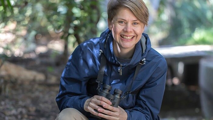 Ann smiles for a photo while crouching down in the forest with her binoculers. 