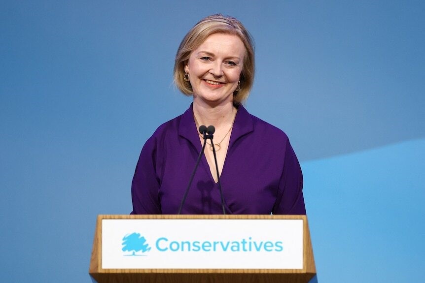 liz truss smiles behind a microphone on a lectern with a banner that says 'conservatives' on it
