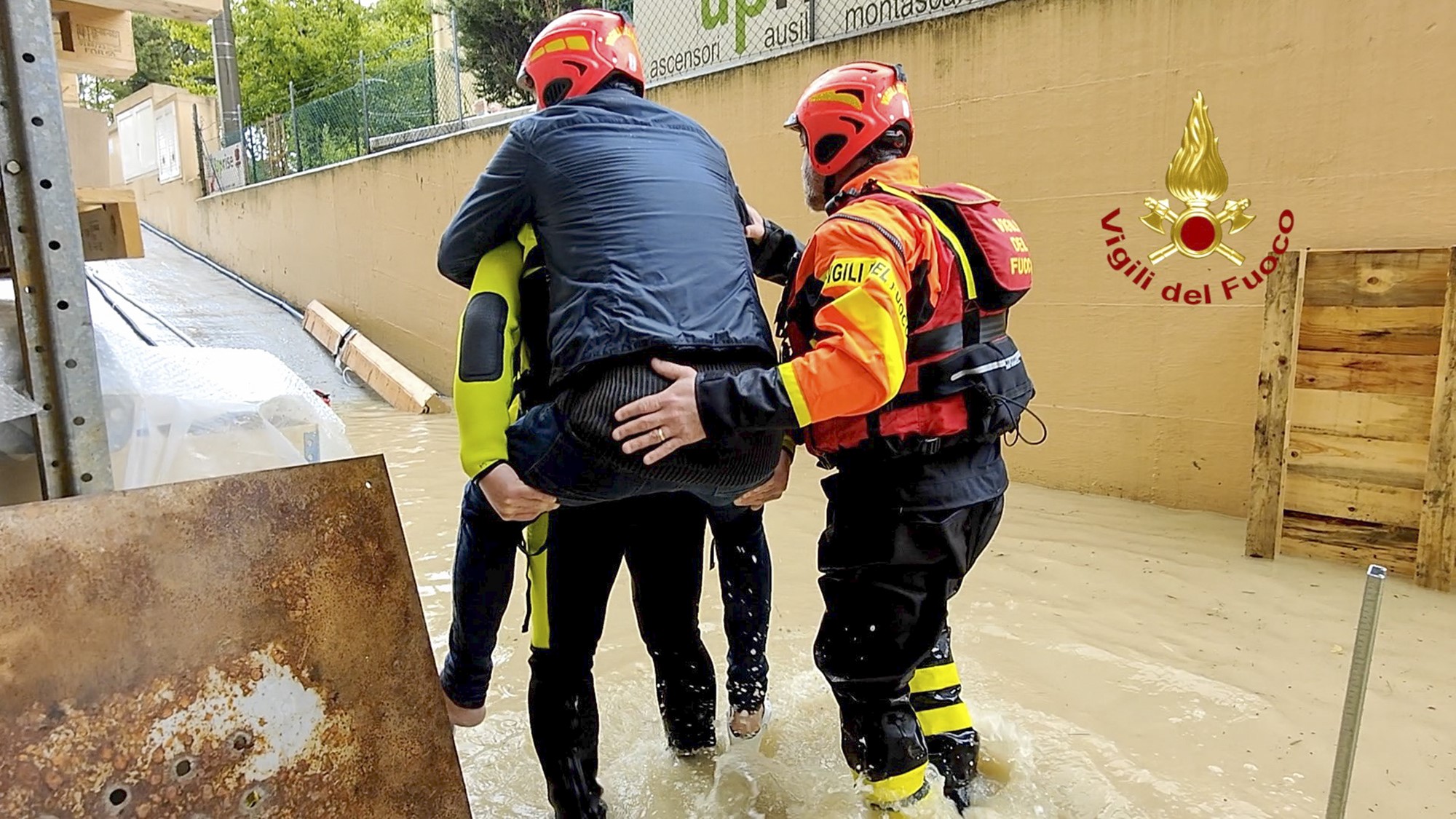 A person carrying another on their back through flood waters, wearing helmets. Another man stands nearby helping.