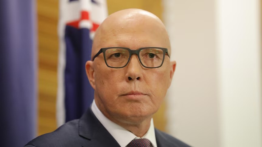 A bald man in glasses and a suit stands in front of an Australian flag