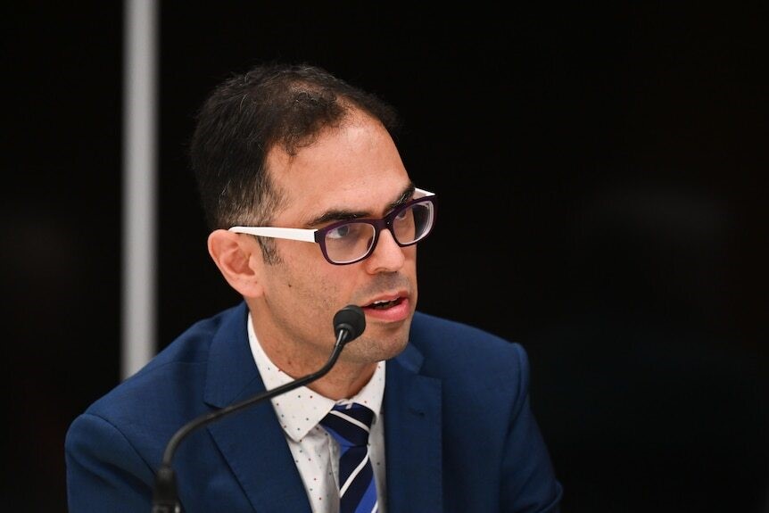 A man with glasses sits at a microphone