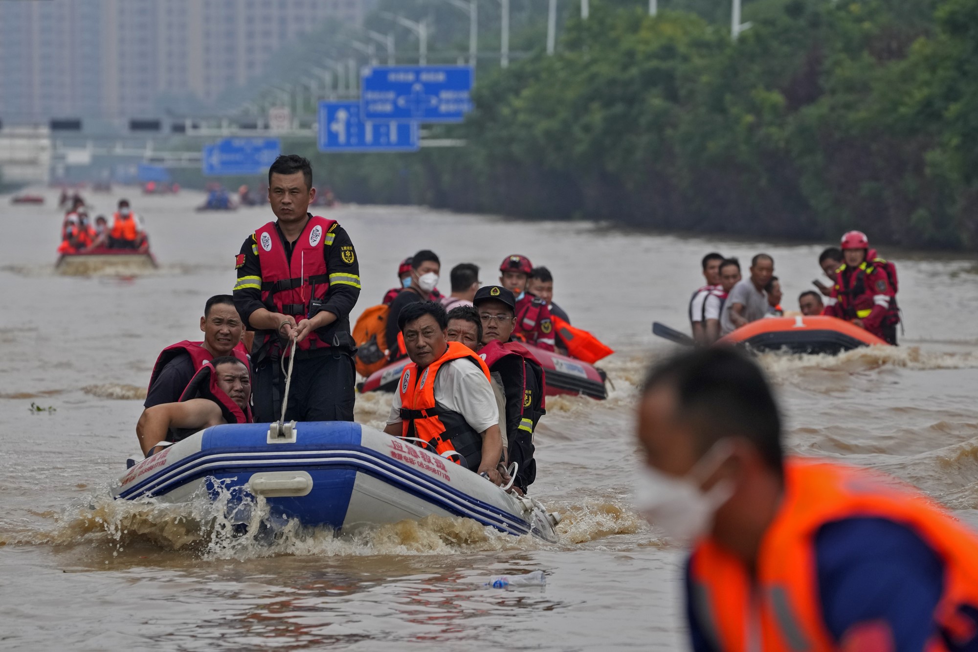 People on small rubber boats moving throug a flooded highway, with trees on the side and tall buildings in the background