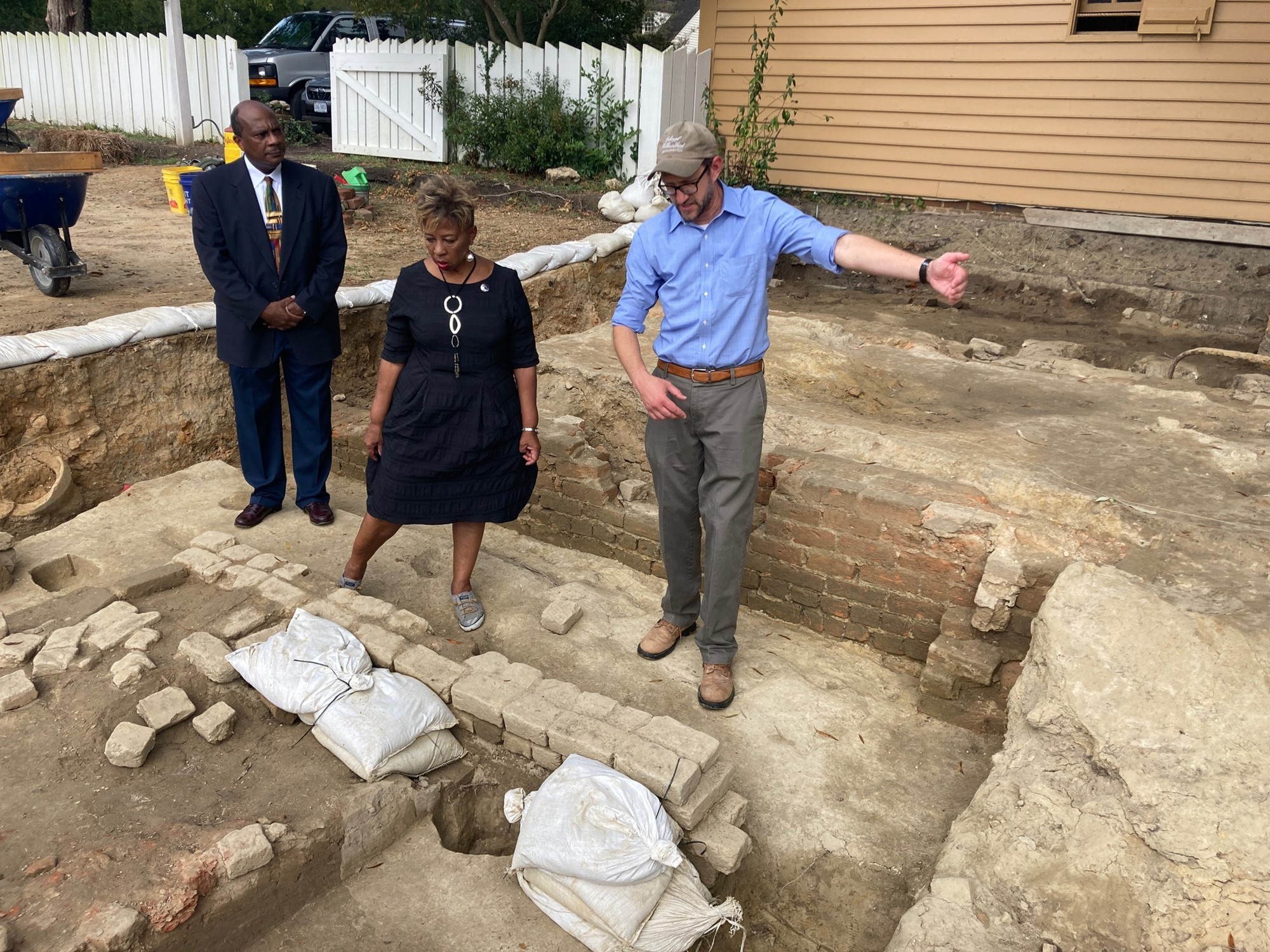 A white man and a black man and woman stand in the excavated remains of a brick building.