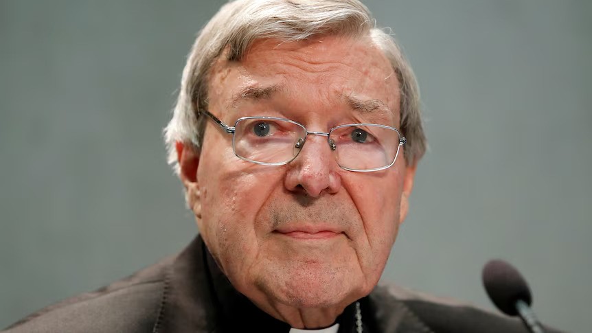 An old man wearing glasses and a priest outfit sits behind a small microphone.