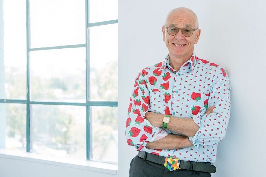 Dr Karl smiles for a photo as he leans against a white wall while wearing a blue and red strawberry button-down.