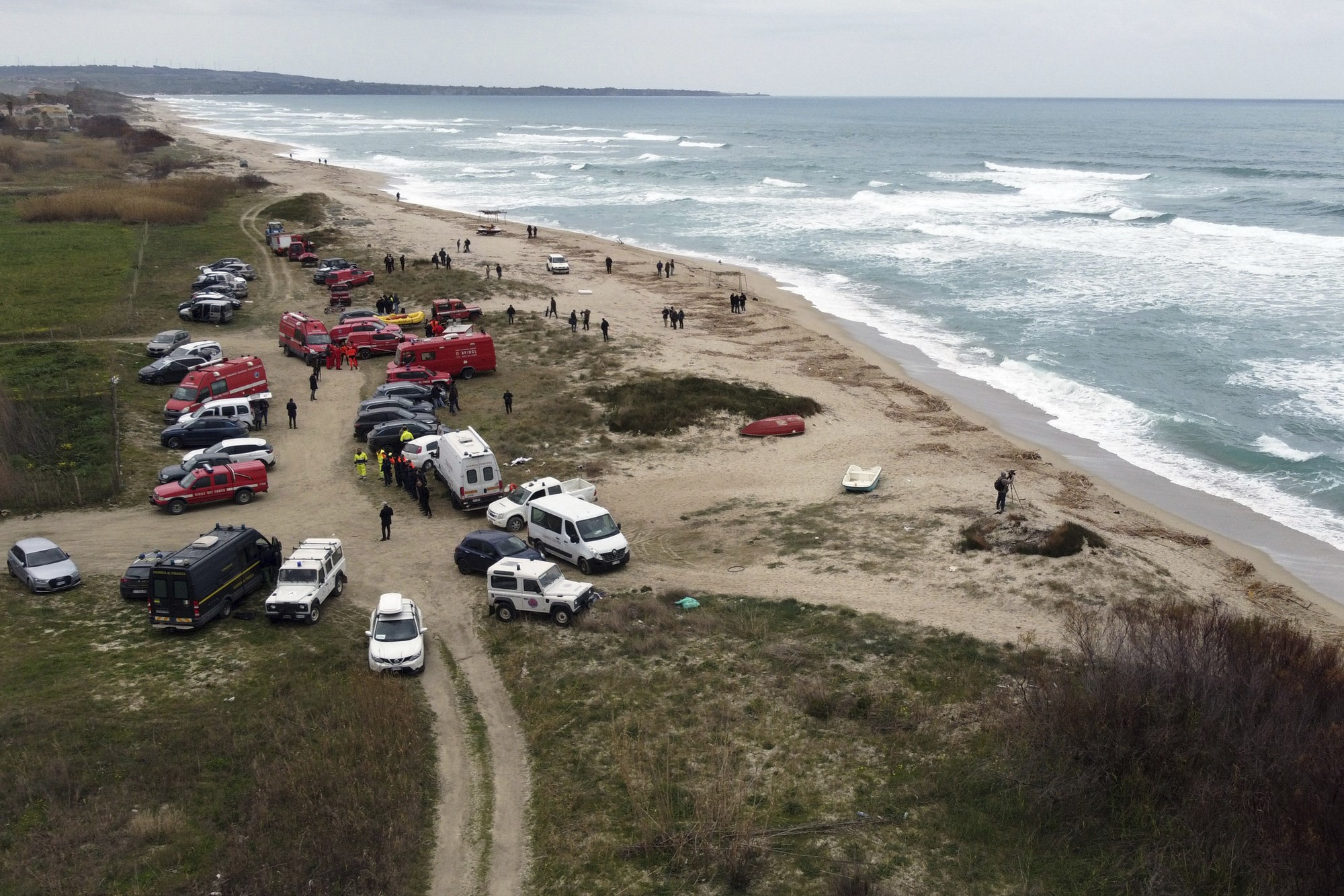 Aerial shot of cars, police and ambulances parked by the side of a beach as people walk over the sand. 