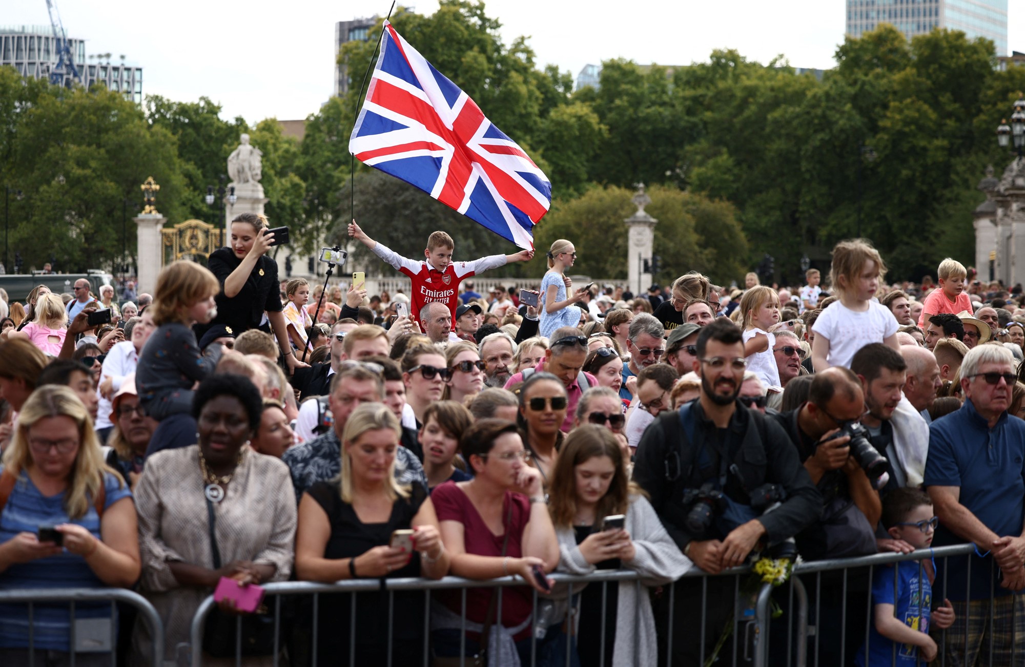 A crowd of people is waiting for King Charles III to arrive. A young boy is waving the British flag above the crowd.