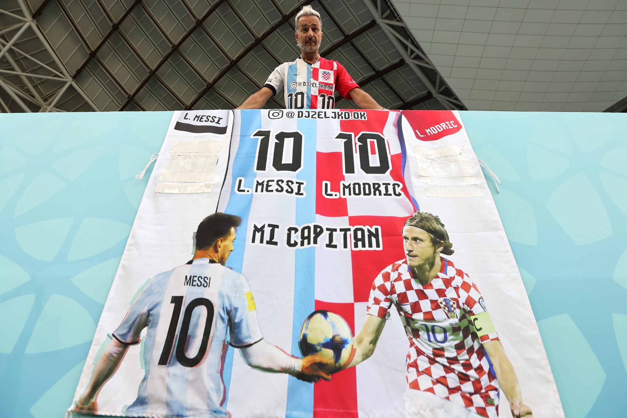A fan wearing a half-Croatian, half-Argentinian jersey stands above a banner with Lionel Messi and Luka Modric on it.