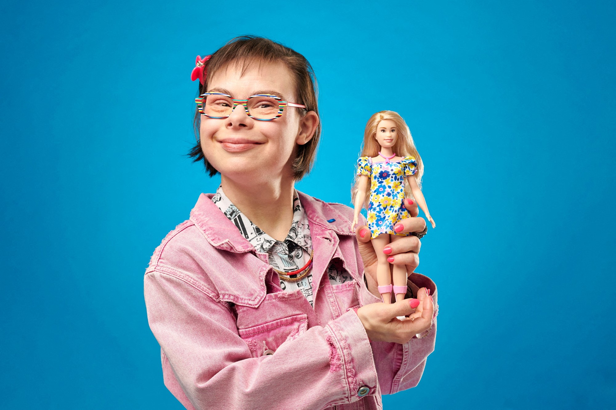 A woman smiles and holds up a barbie in front of a blue background. 