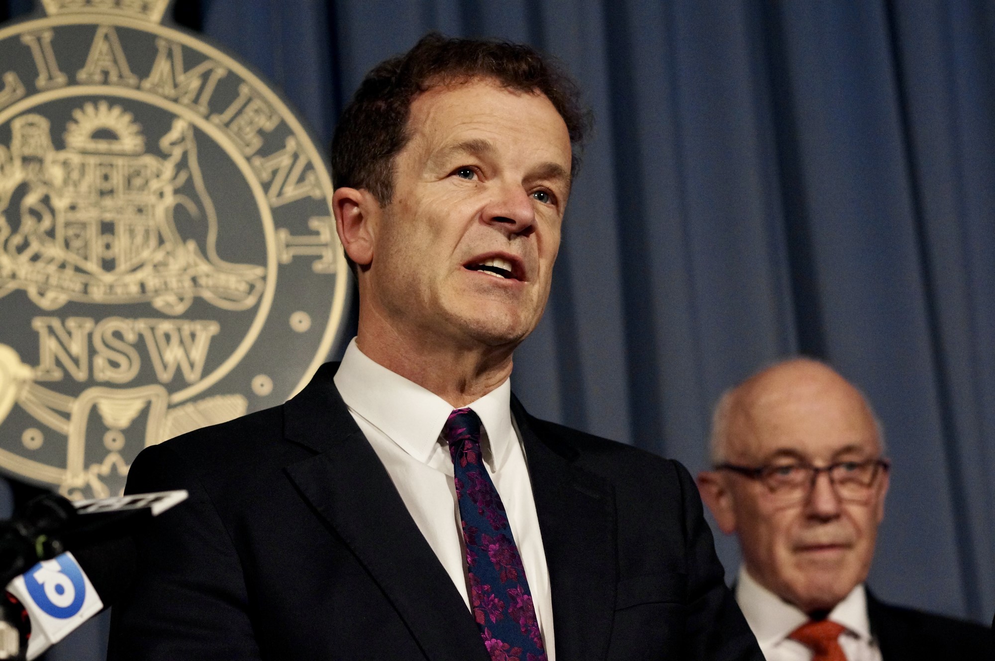 A man in a suit and tie speaks in front of a paliament of nsw symbol