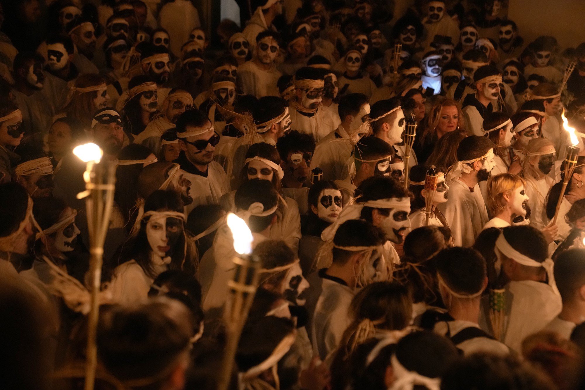 A crowd of people all with faces painted in black and white and wearing white robes walk through the street, some holding torches. 