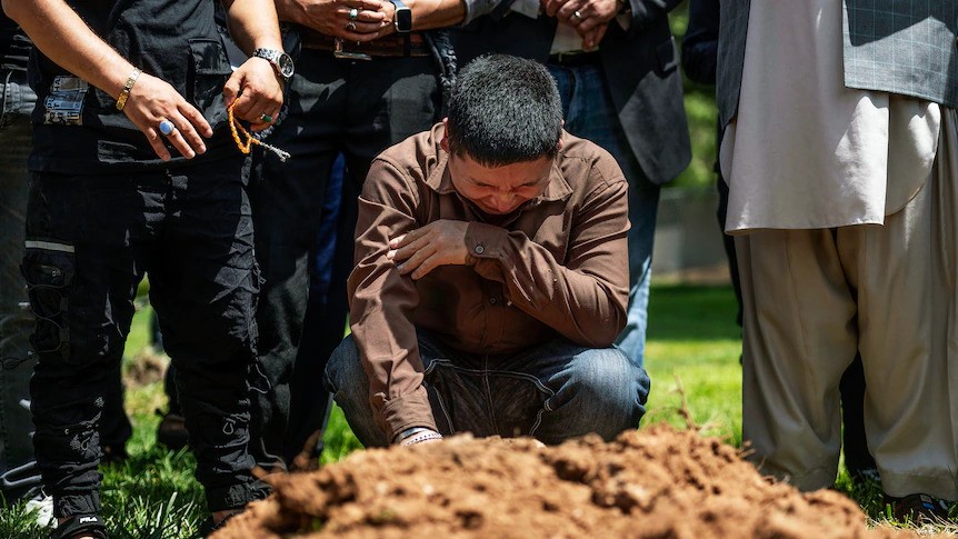 A man cries while kneeling near a pile of dirt (a grave), as others stand around him