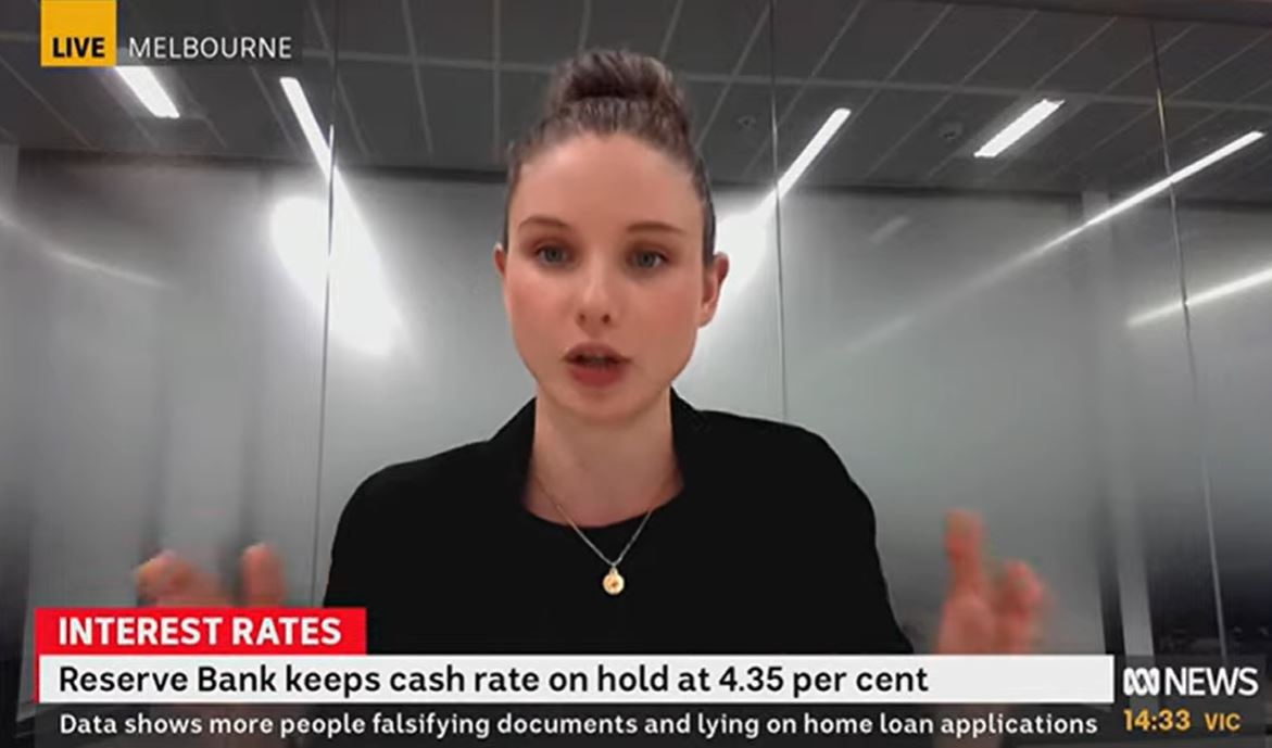 A young woman with her hair in a bun wearing a black shirt and jacket sits in a glass office gesturing with her hands and talking. Text explaining the latest interest rate decision by the RBA is on the bottom of the screen.