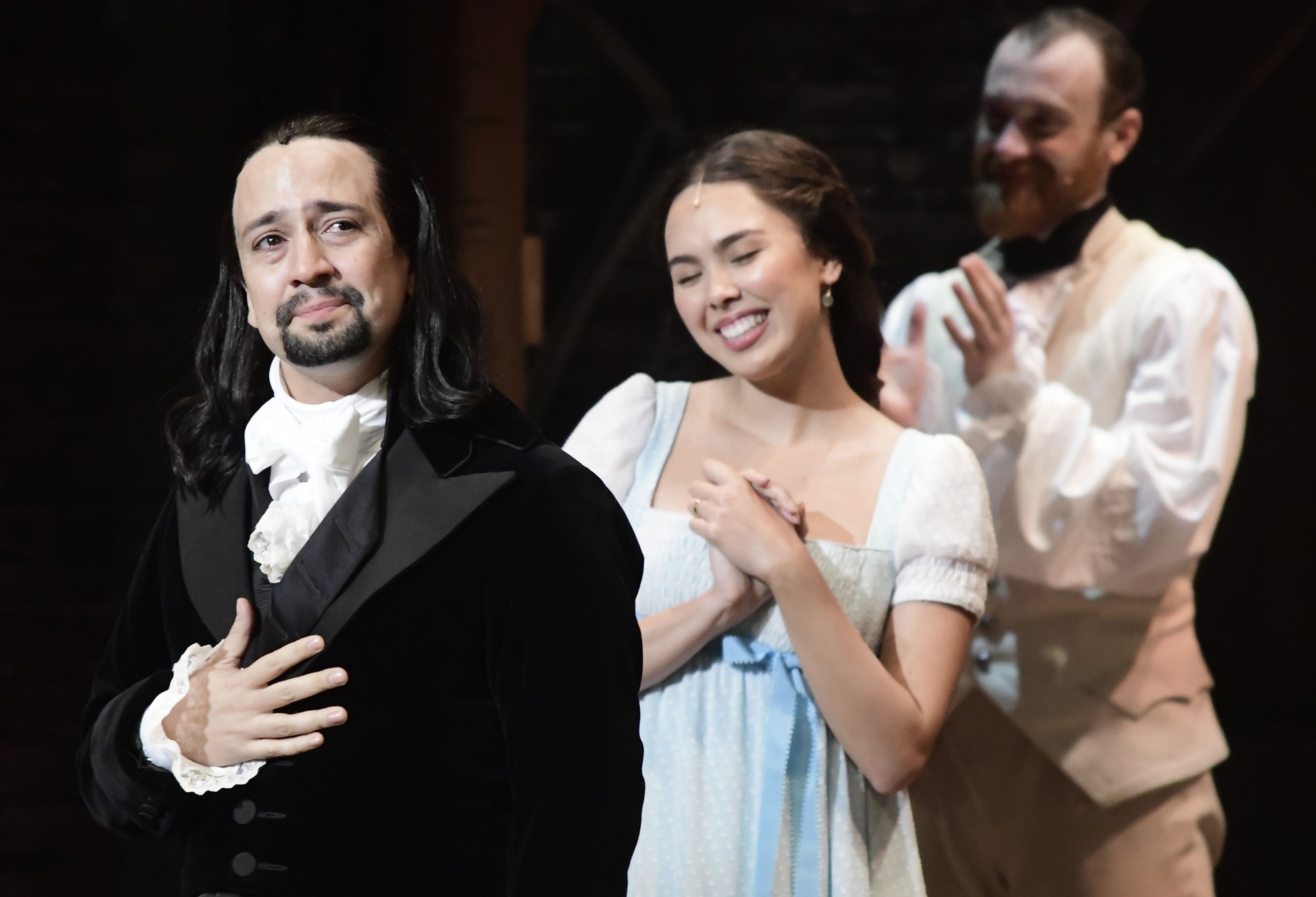 Lin-Manuel Miranda in Hamilton costume stands with hand on chest as two other actors clap
