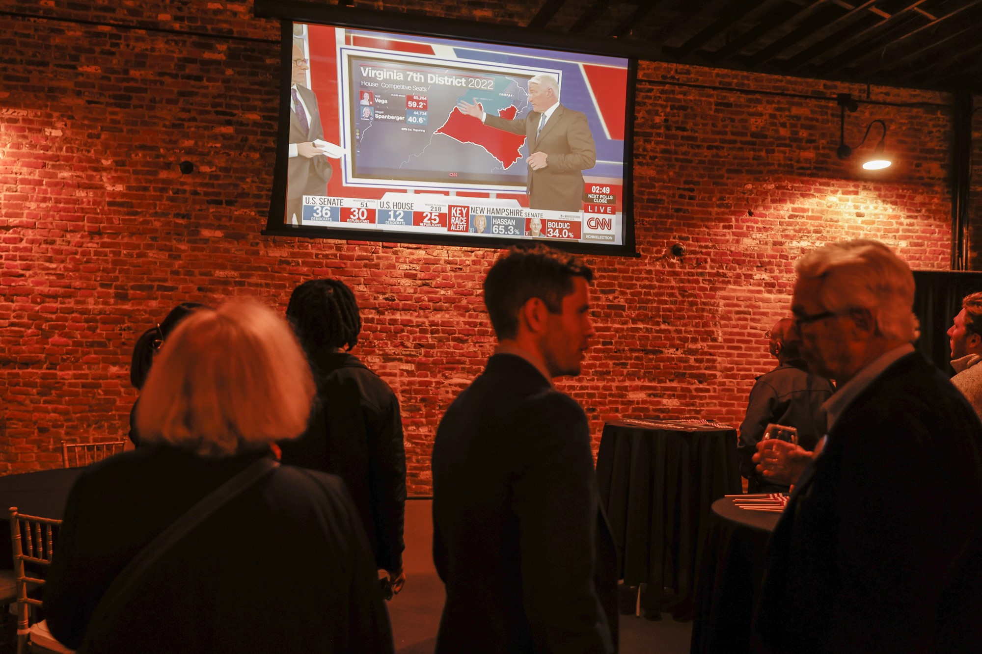 People stand around with drinks in a venue while a TV shows the midterms coverage.