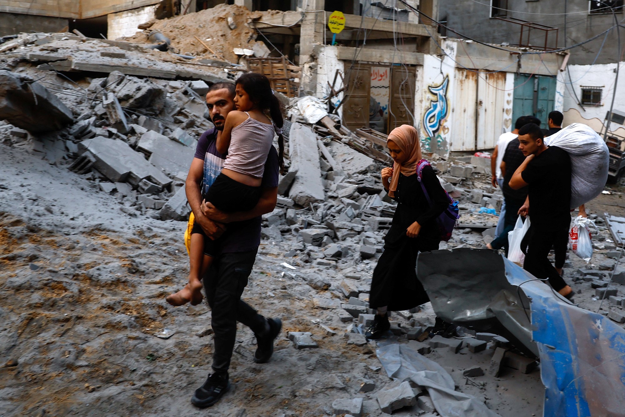 A man carrying a child and two women walk through a destroyed street