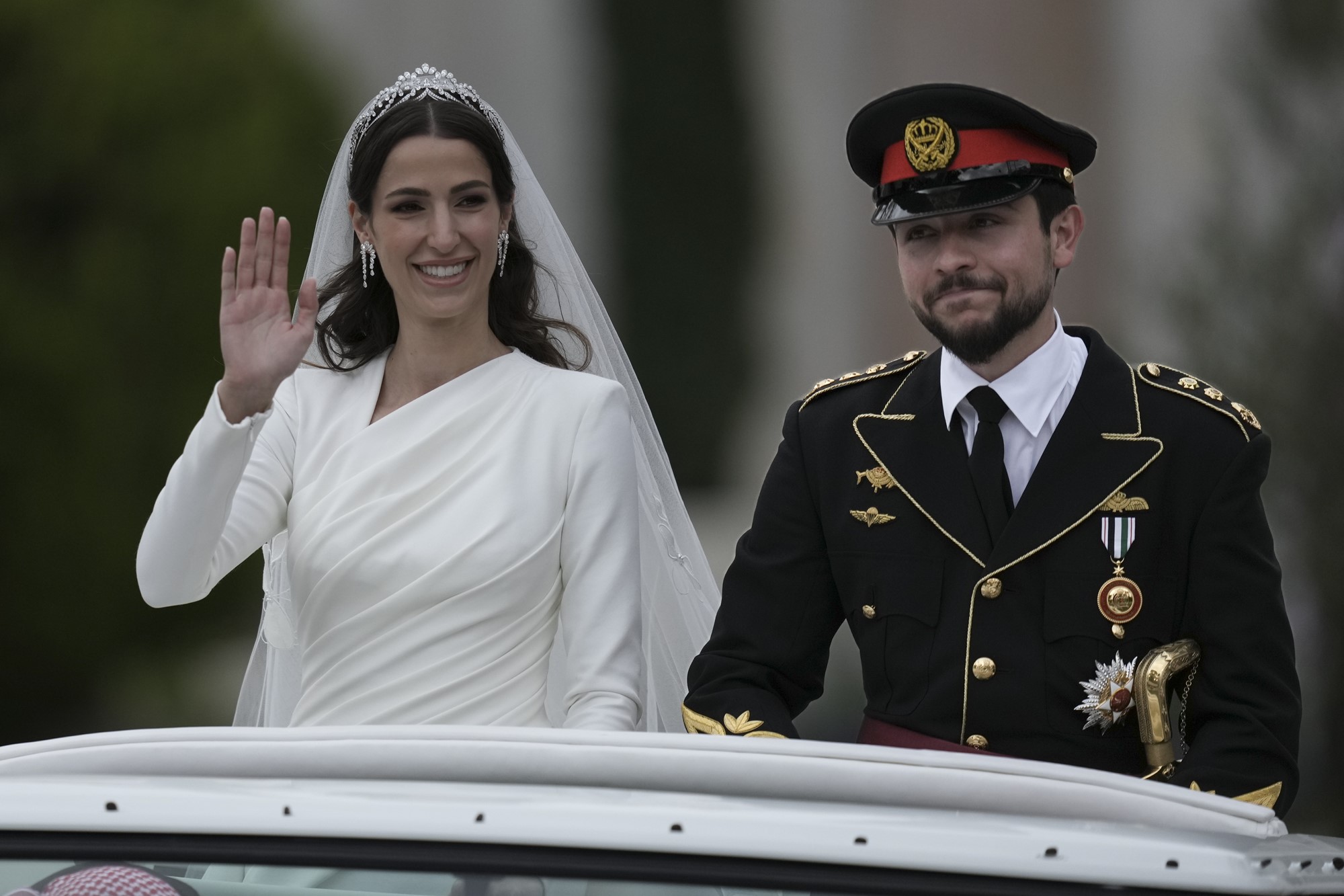A bride in a white dress and veil, and a groom in a military outfit, smile and wave standing out the top of a moving vehicle