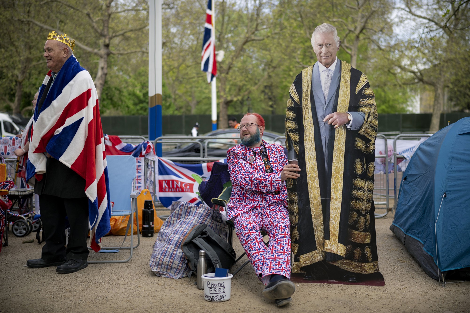 A man in a Union Jack suit sits in a chair near a fence next to a road, with a cardboard cutout of a standing Kind Charles