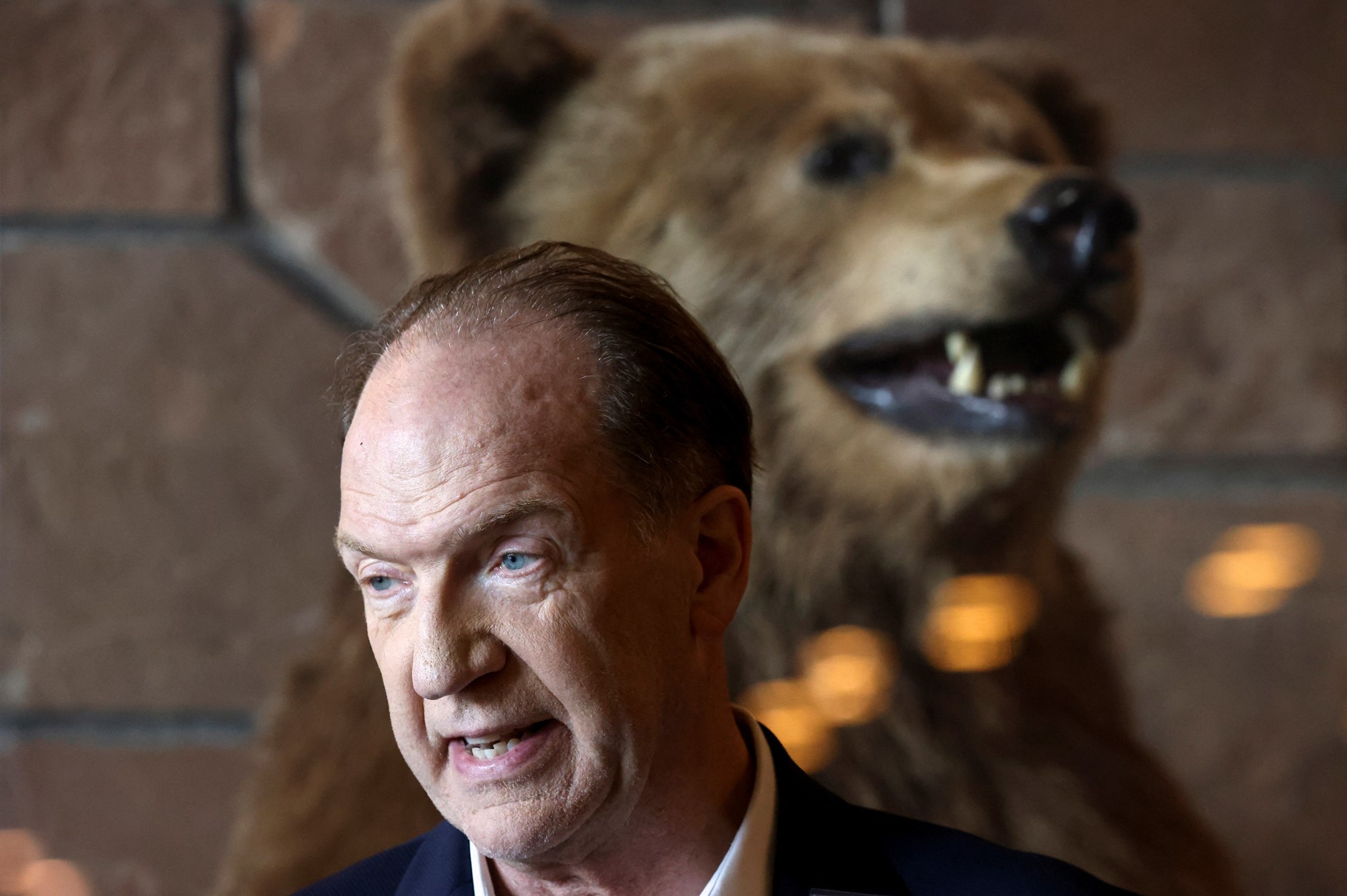 An in-focus shot of an older man with a taxidermied grizzly bear in the background.