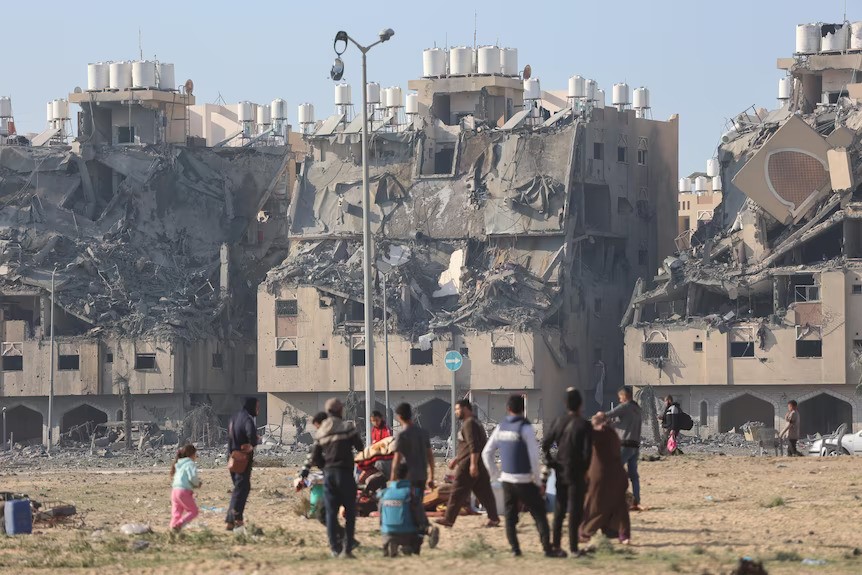 Buildings in ruin in the background as people stand around in the foreground