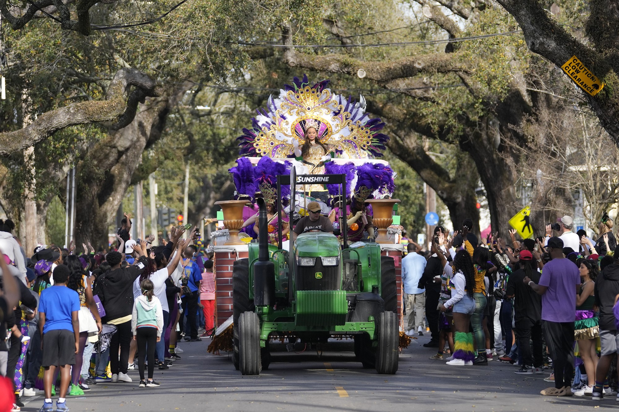 A woman in elaborate costume sits on the back of a parade float pulled by a tractor through the street. 