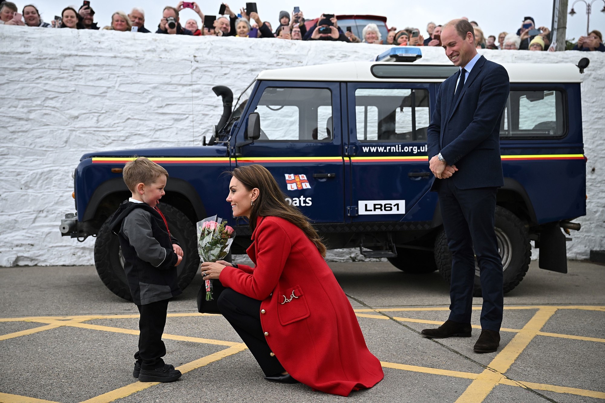 Princess Catherine crouches to greet a young boy. Prince William stands behind Catherine