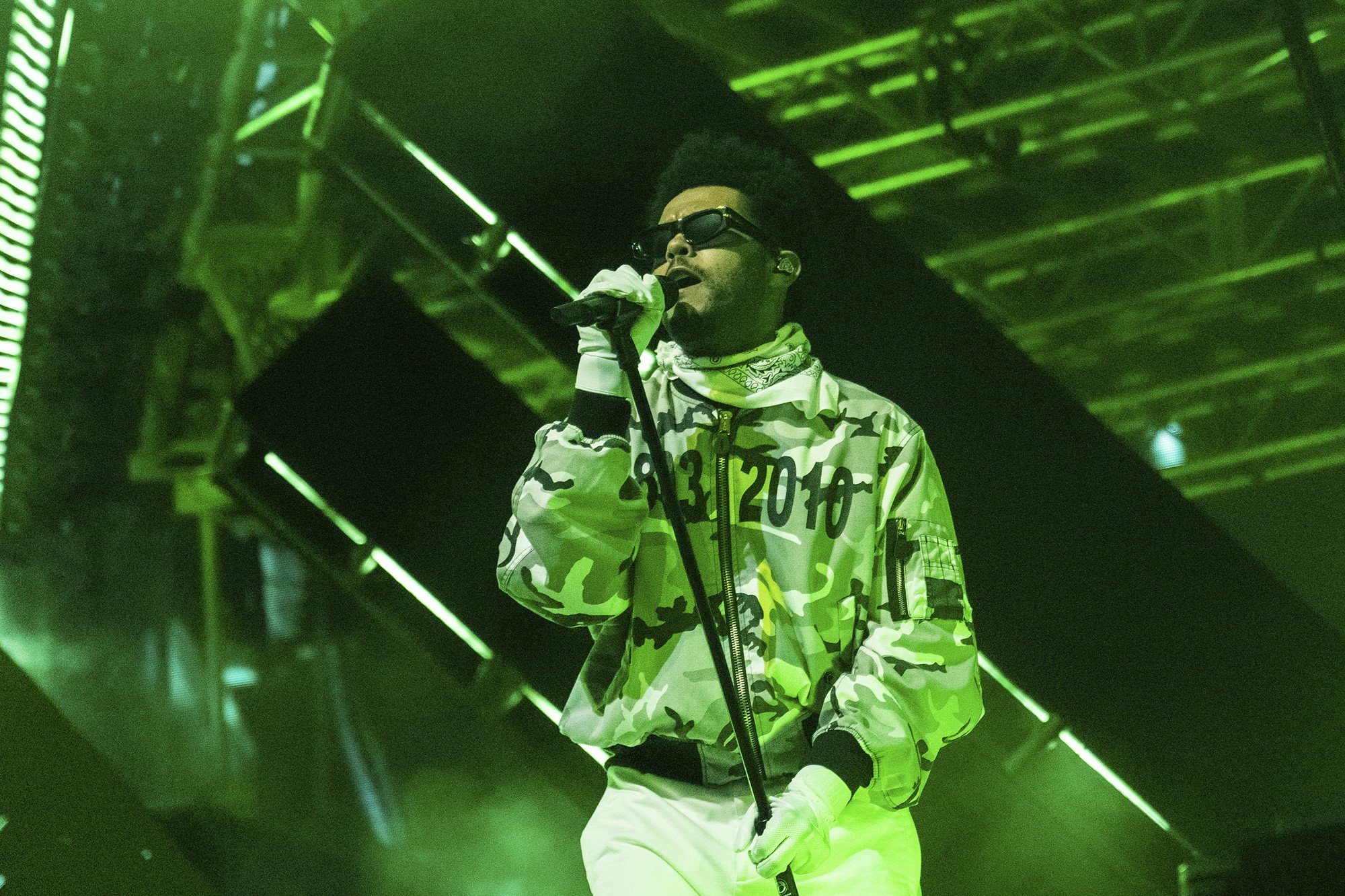 The Weeknd sings on stage, wearing a camo jacket, sunglasses and gloves