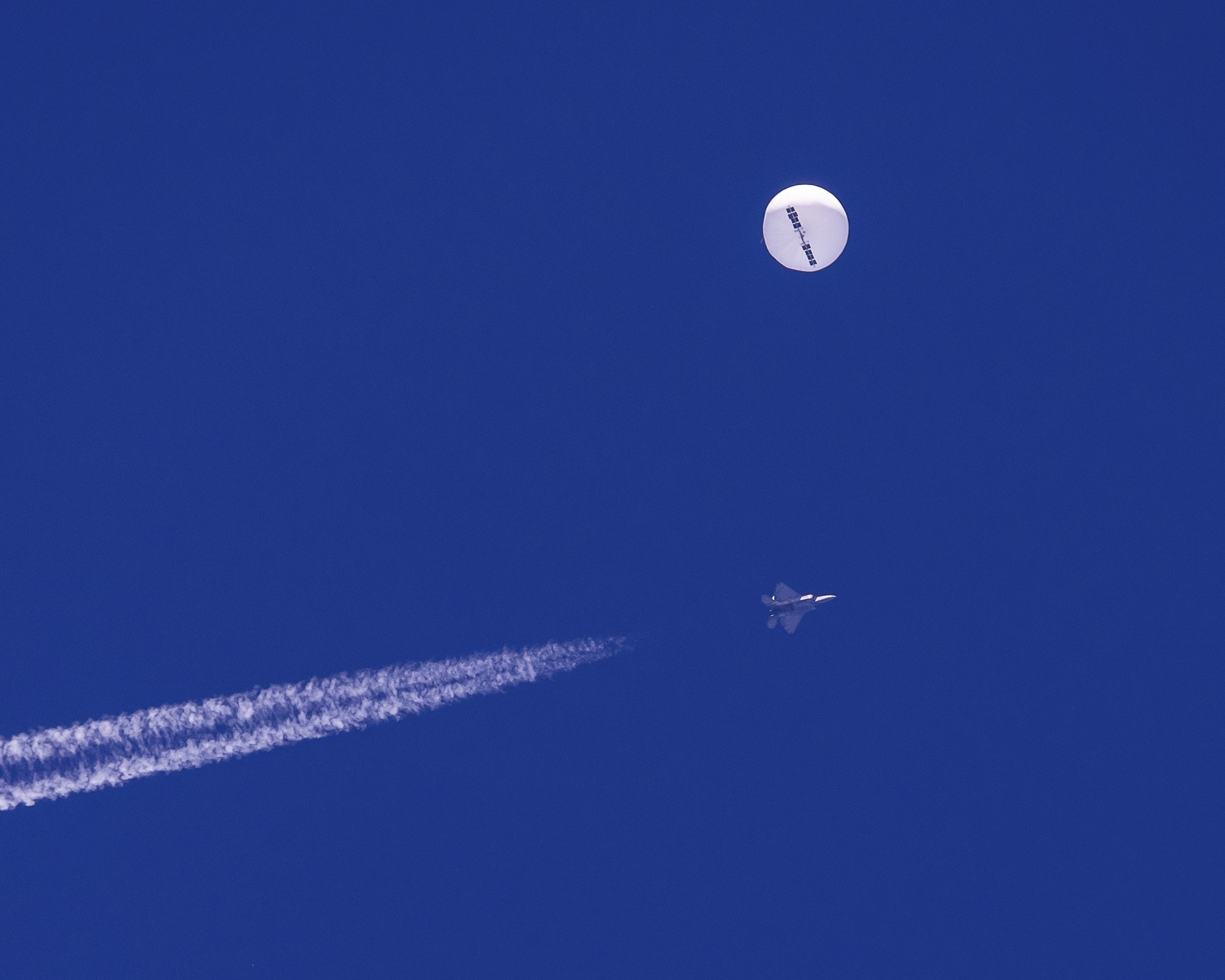 A balloon floats in the sky as a jet flies past leaving a trail of smoke. 