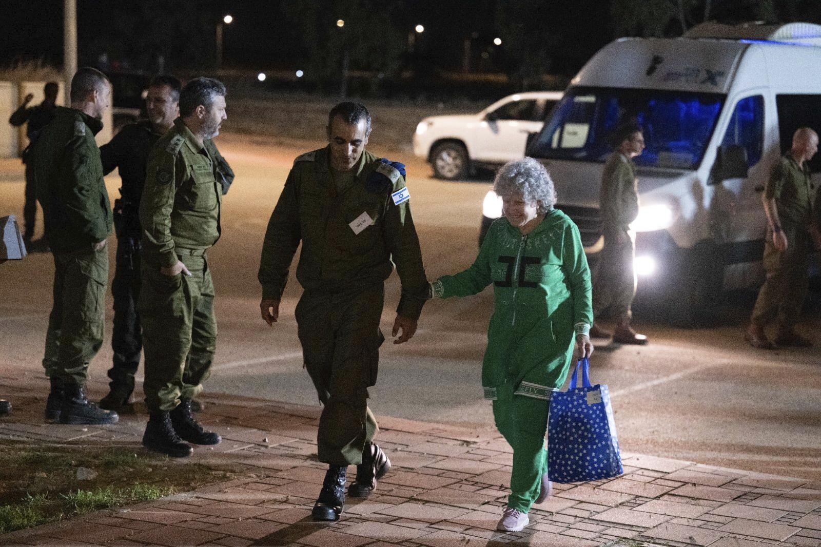 An elderly lady walks next to an Israeli soldier at night, holding his arm. A van and other soldiers are behind them