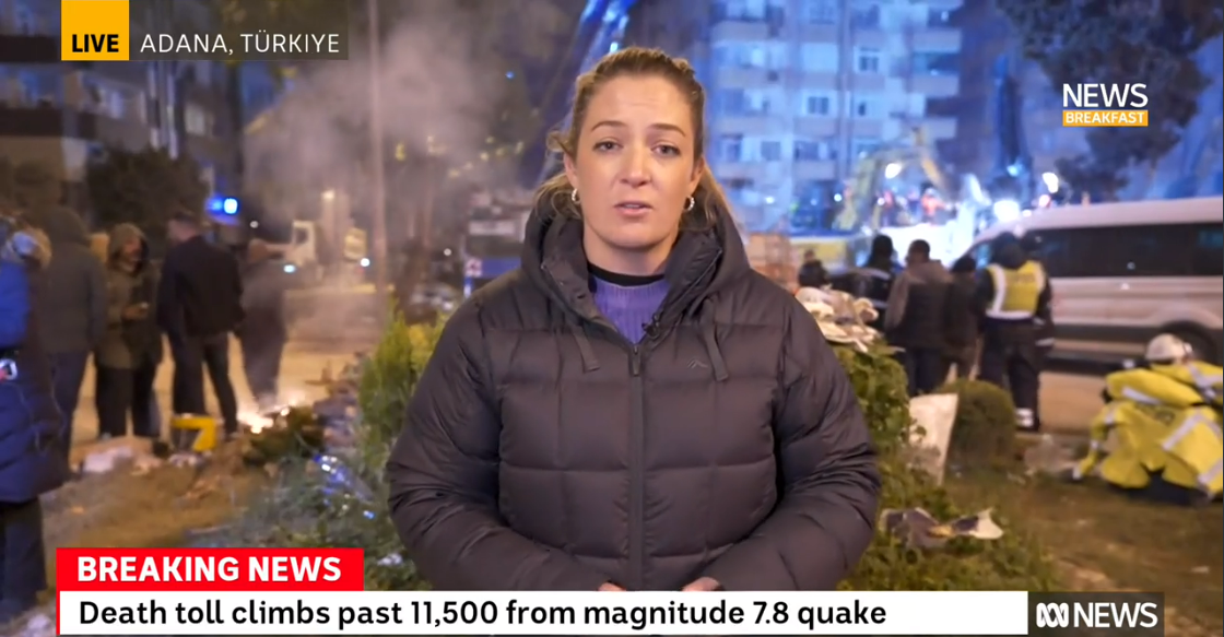 A female reporter stands and speaks near a destroyed building, with other people nearby