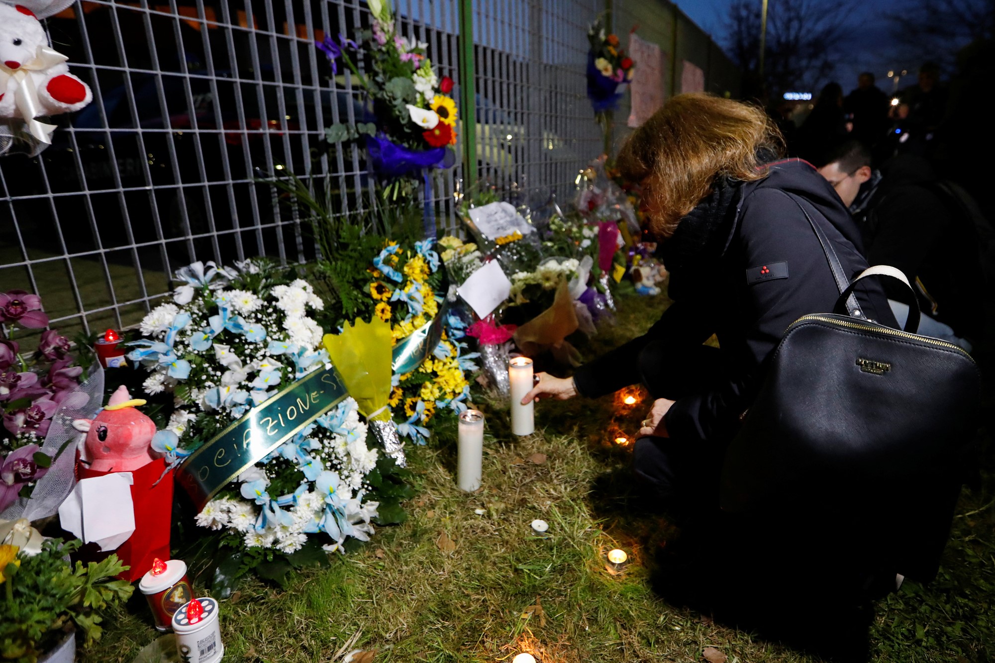 Awoman kneels to place a candle next to a metal fence next to bouquets of flowers and toys. 