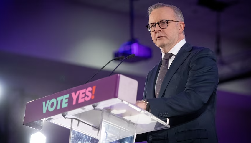 A middle-aged man in a suit with grey hair and wearing glasses stands behind a lectern.