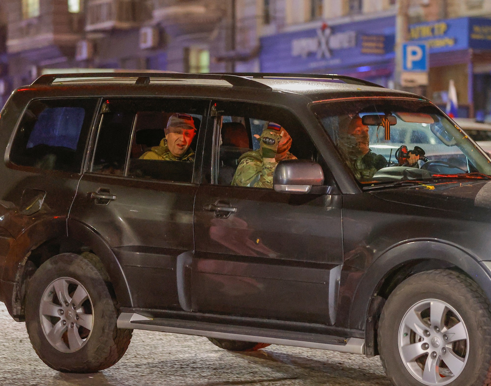 Yevgeny Prigozhin sits in the back of a car with two people in the front of the car in military gear.