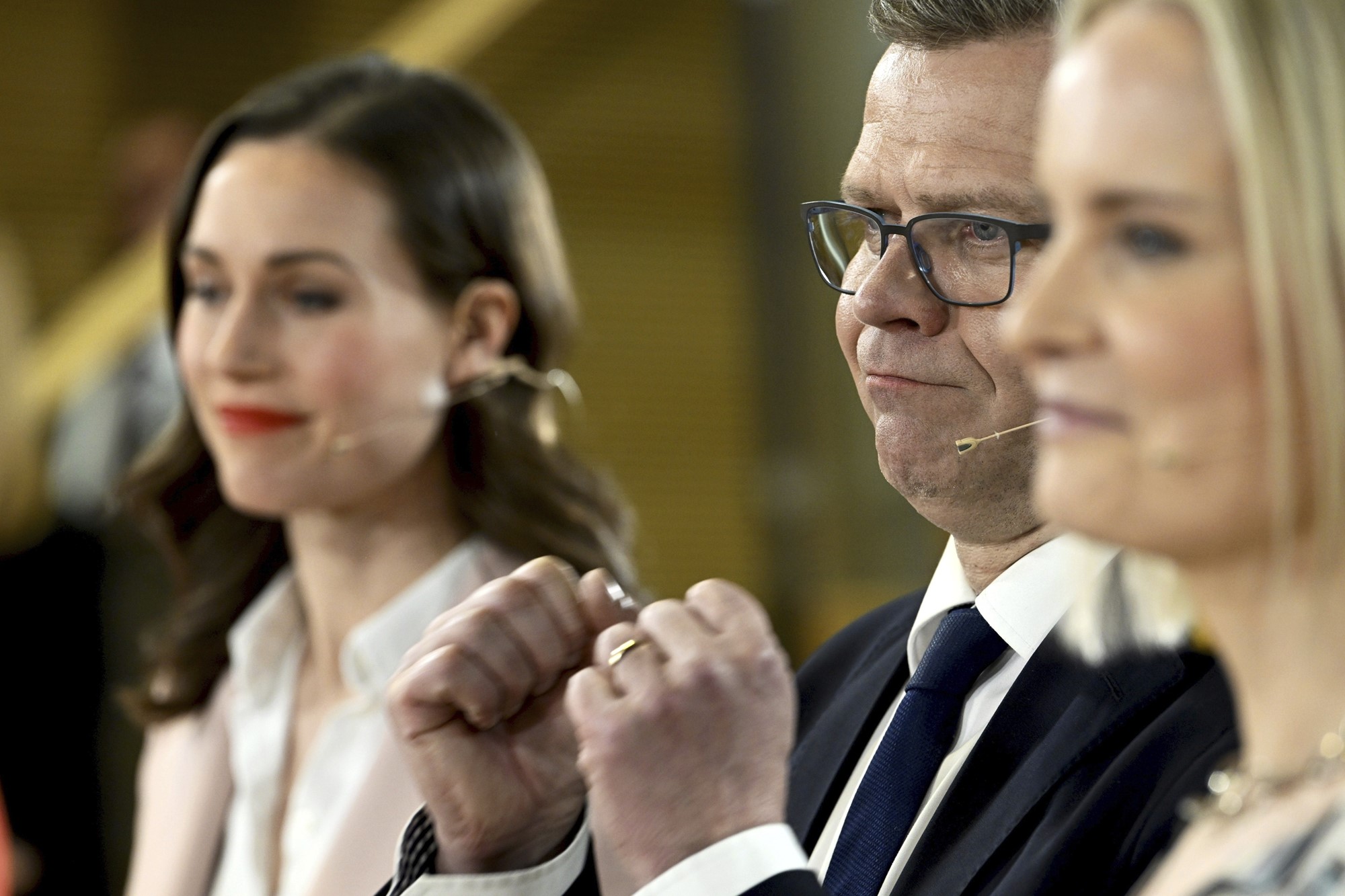 A white man with glasses and a suit clenches his fists. Two women are standing either side of him