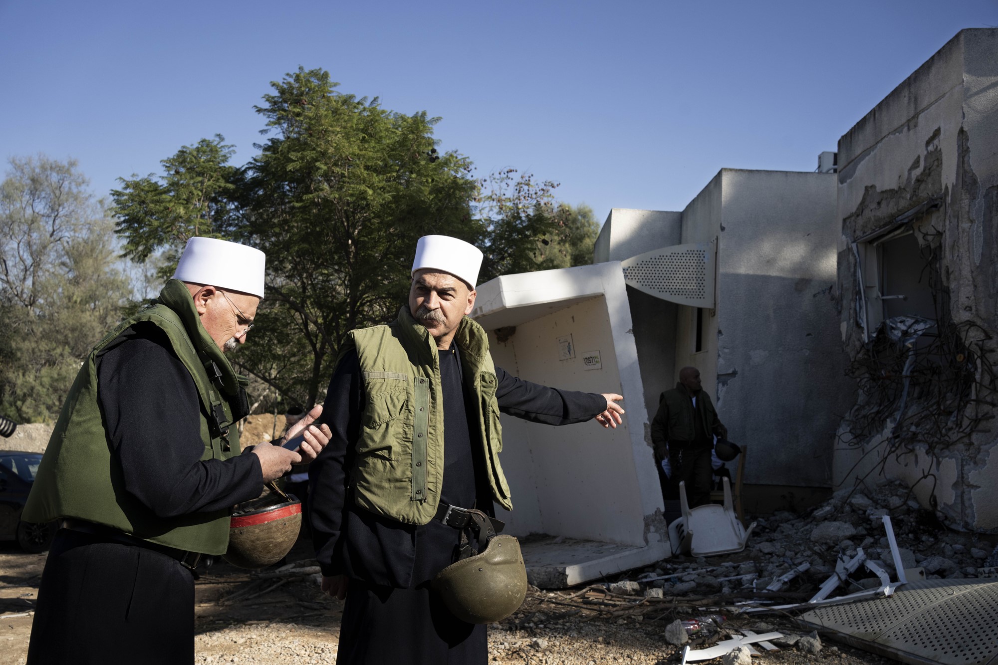 Two Druze faith leaders in traditional headwear stand near a damaged building, both carrying helmets. One points to the building
