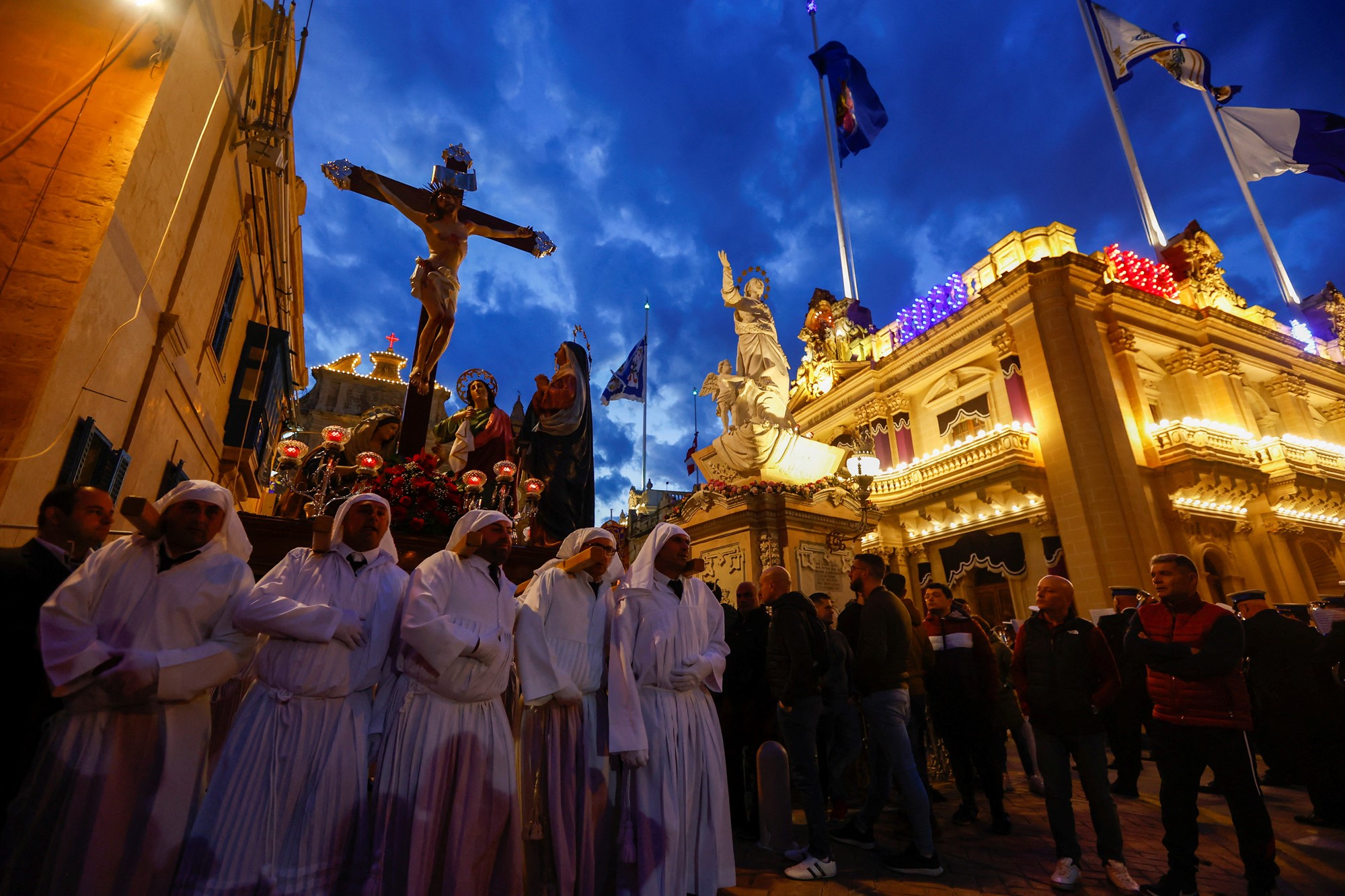 Crowds of people in hooded white robes stand outside under a statue of Christ on the cross. 