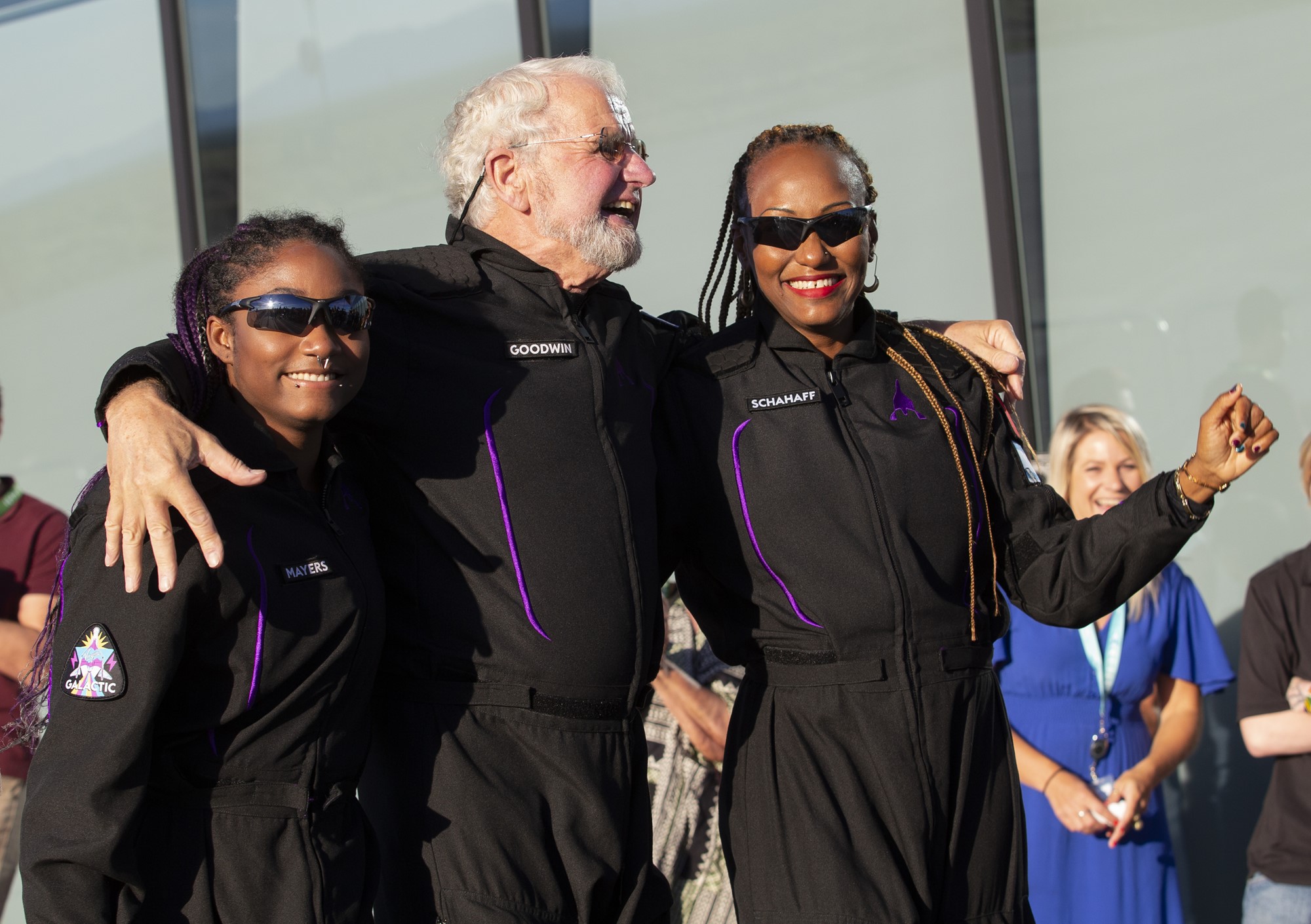 Two black women with braids and sunglasses in space suits stand either side of an elderly white man with a beard