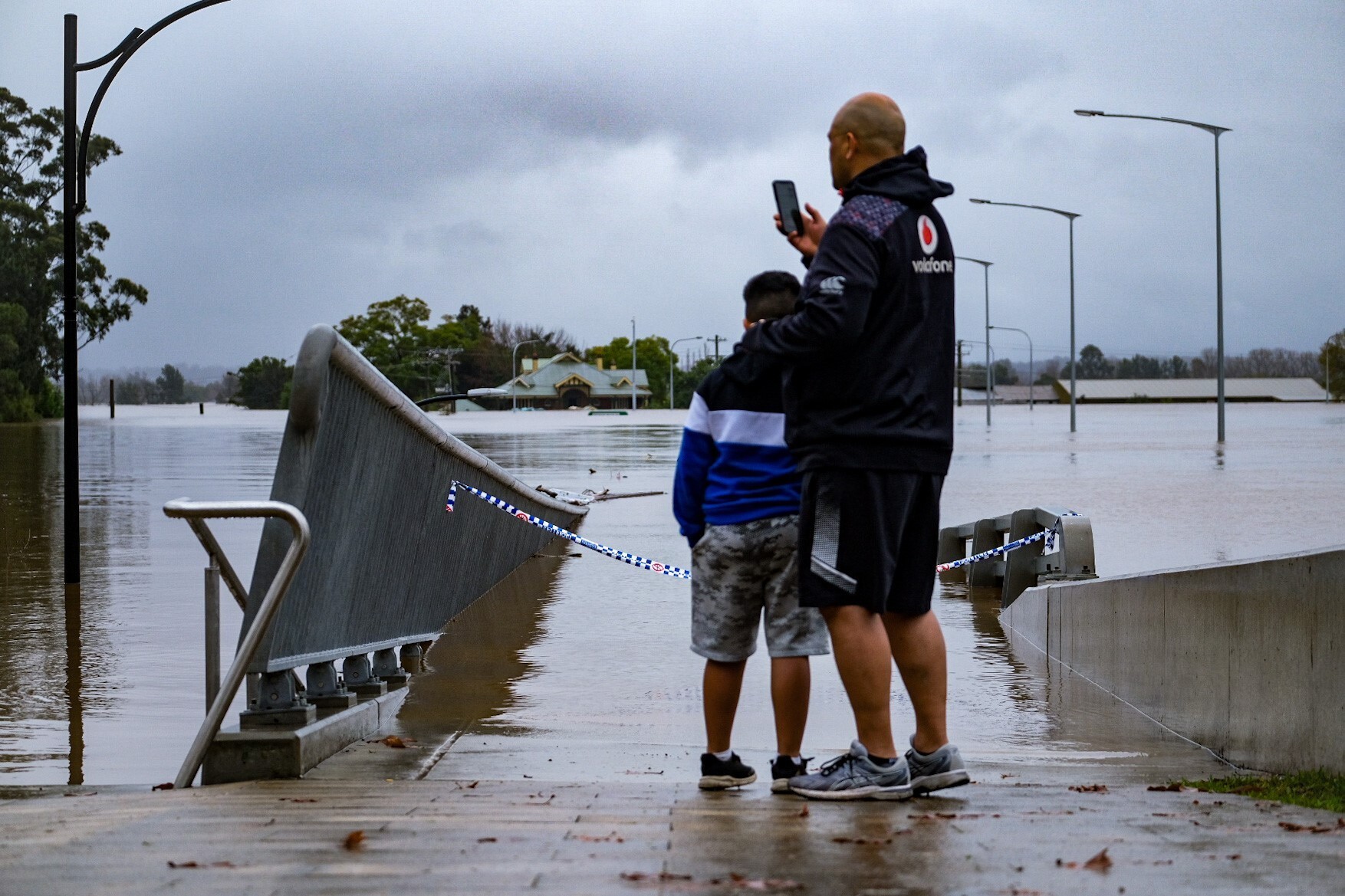 a man stands at the edge of a flooded walking bridge with his arm around a child taking a photo on his phone