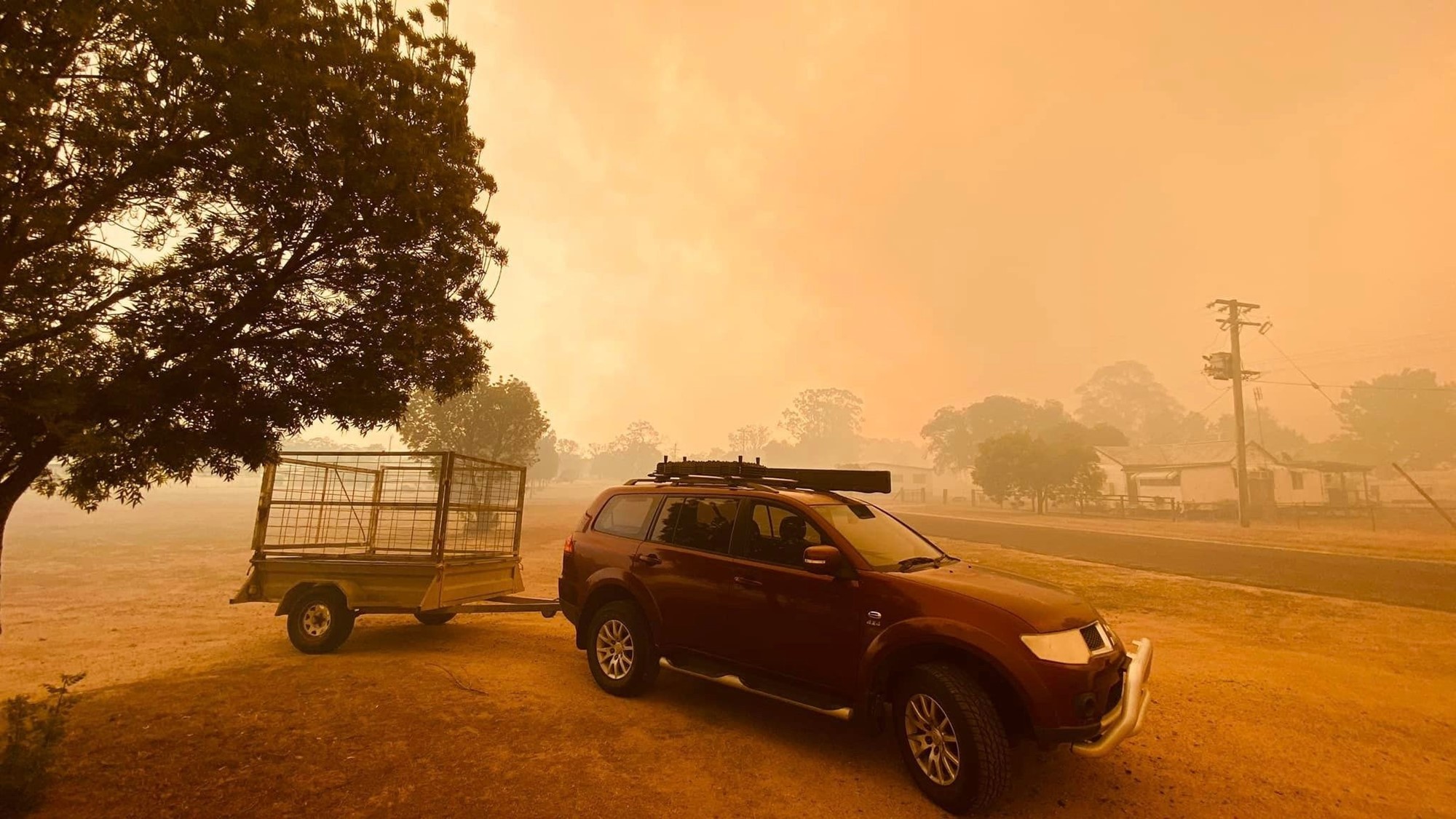 a car with a trailer parked under a tree beside a rural road, the sky is orange and there is bushfire smoke everywhere