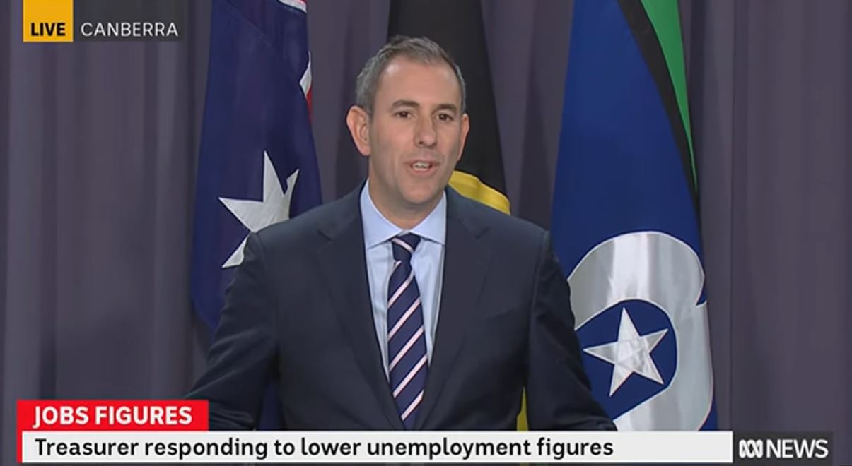 A man wearing a suit stands in front of an Australian, Aboriginal and Torres Strait Island flag.