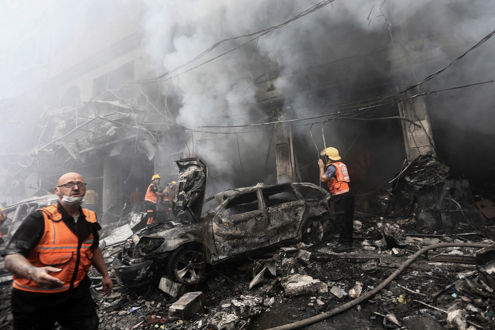 People in high vis vests walk around the remains of a burnt out car and building