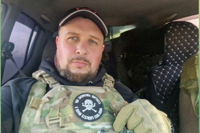 A man taking a selfie in a car, wearing a hat, hoodie and military jacket and gloves