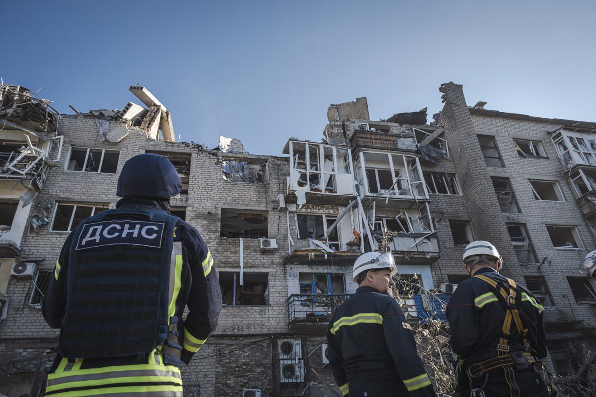 Men in firemen uniforms look up to a residential unit block which has been badly damaged by shelling
