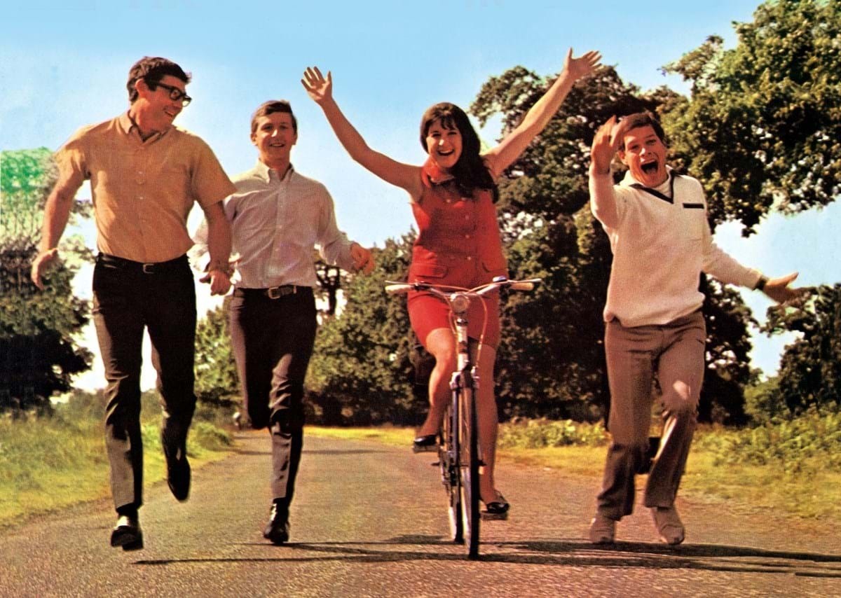 The members of The Seekers run and cycle down a sunlight path bordered by trees.