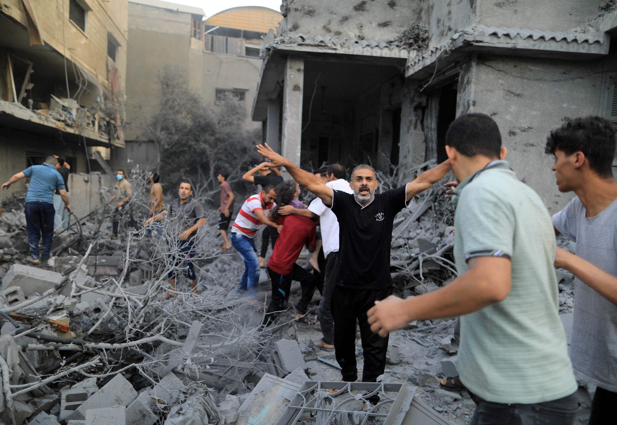 A man stands with arms outstretched as a crowd behind him dig through the rubble of a house