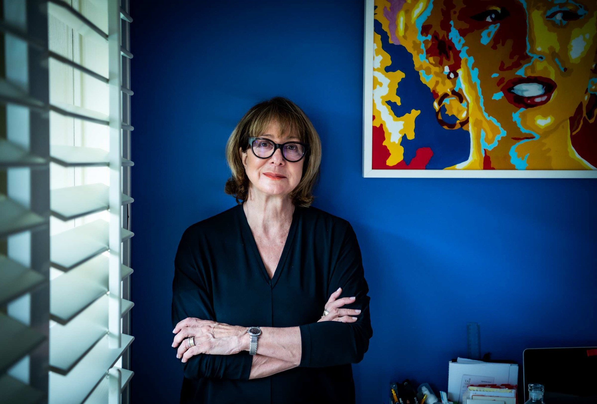 A woman with glasses leans against a dark blue wall