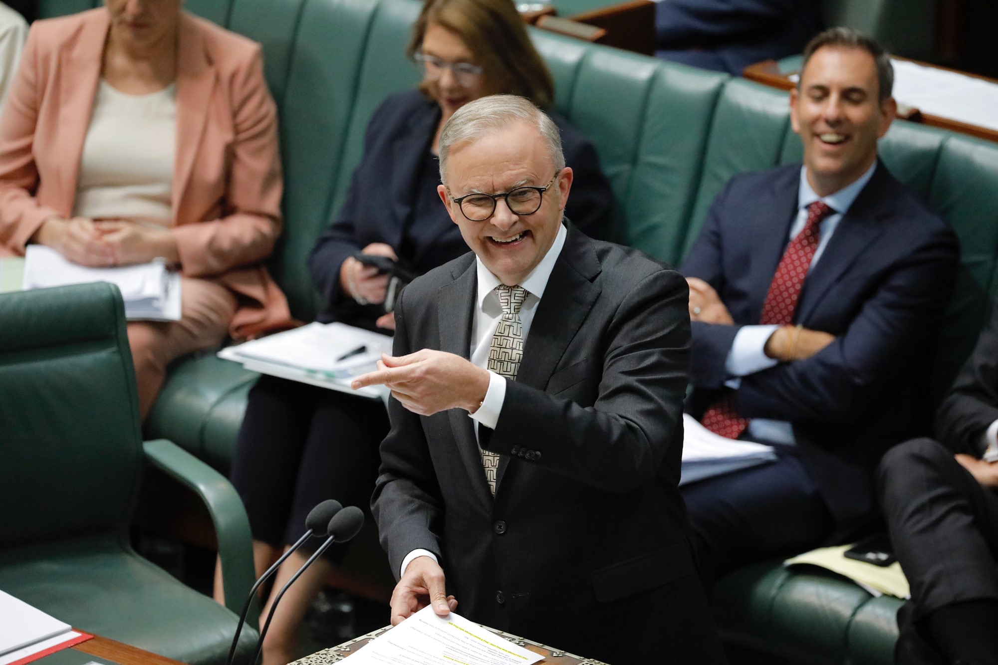 A man in a suit, tie and glasses smiles and points in parliament